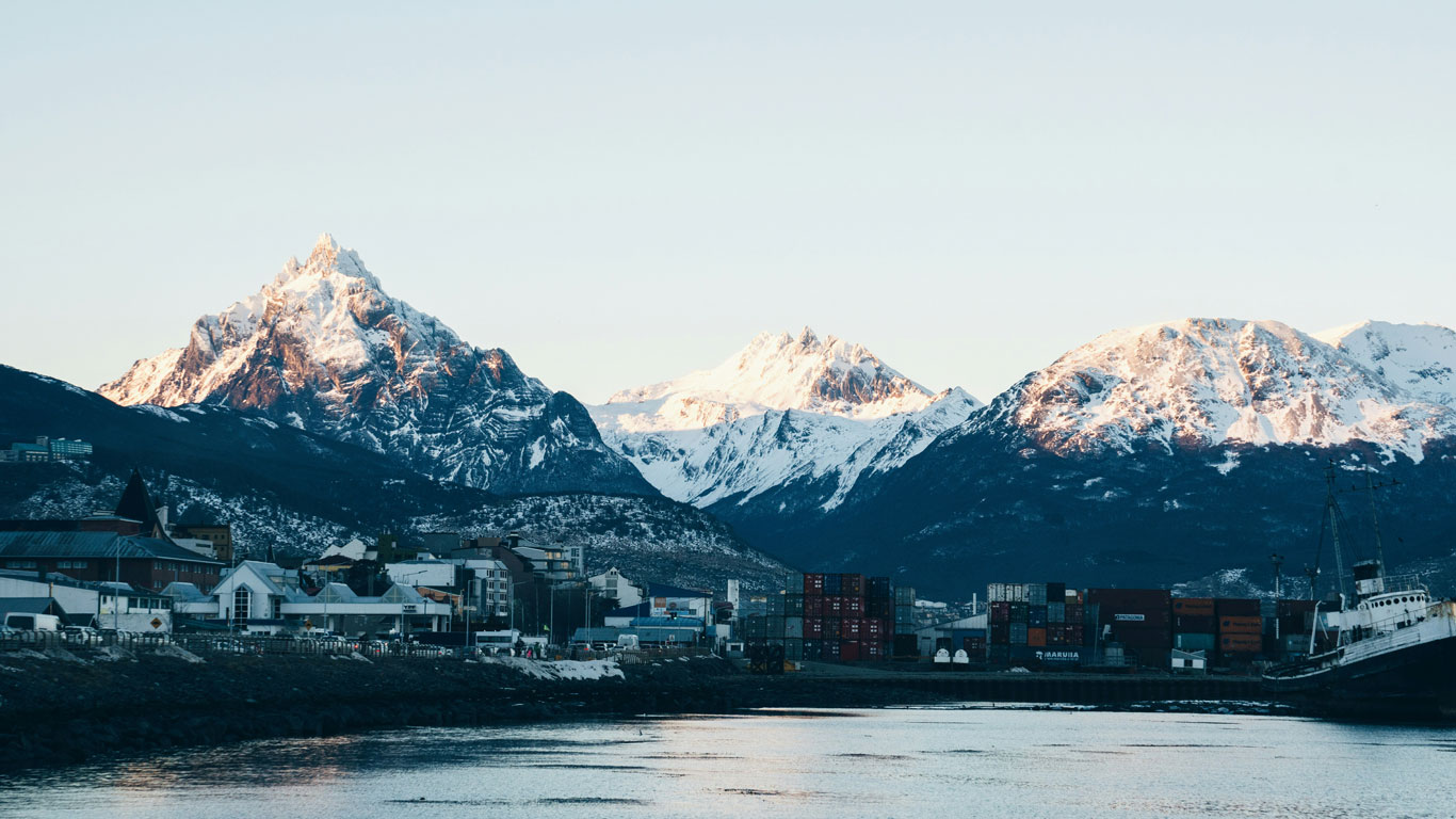 Vista de la ciudad de Ushuaia con el puerto y contenedores junto a un barco, con montañas de picos nevados al fondo iluminadas por la luz del sol.