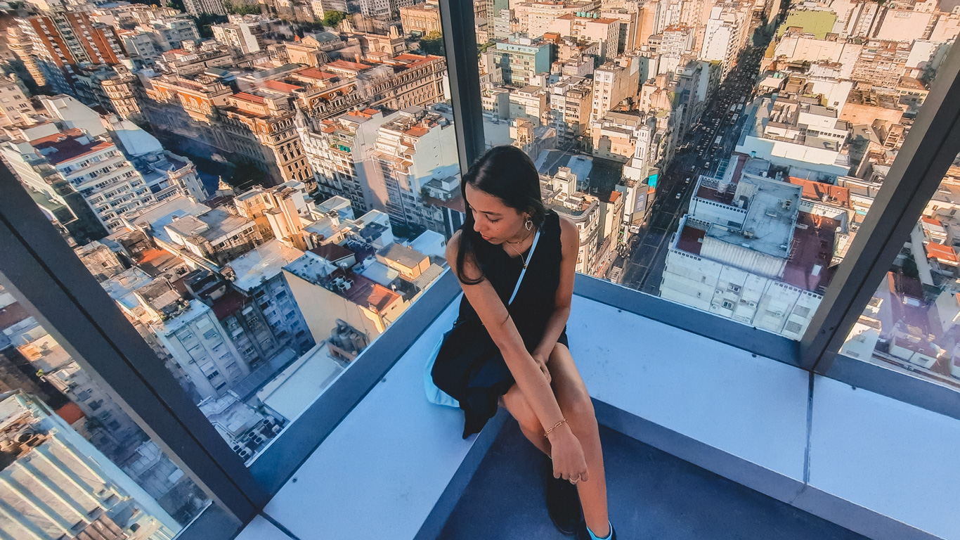 olympo-sky-bar-buenos-aires Una mujer sentada en el mirador acristalado del Olympo Sky Bar, ubicado en lo alto de un edificio, disfruta de una vista panorámica impresionante de la ciudad de Buenos Aires al atardecer.