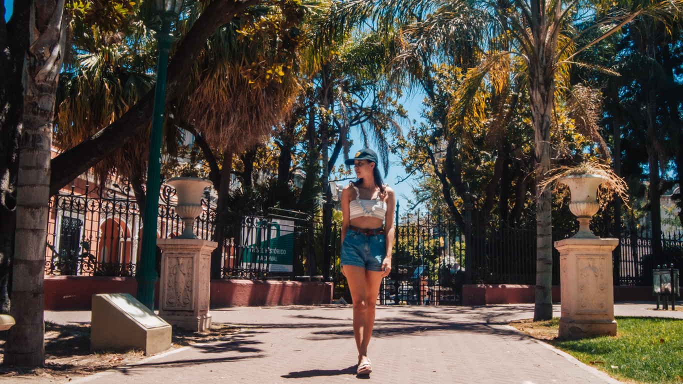 parque-lezama Una chica camina por el Parque Lezama, siguiendo un sendero flanqueado por palmeras y macetas ornamentales frente al Museo Histórico Nacional, bajo un cielo azul intenso.