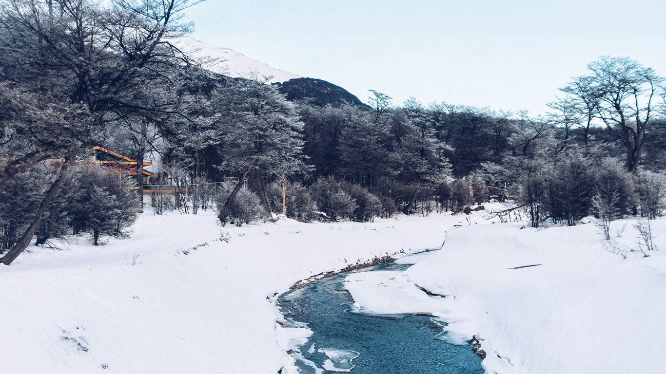 Arroyo parcialmente congelado serpenteando por un bosque cubierto de nieve en el Parque Nacional Tierra del Fuego, con montañas al fondo.
