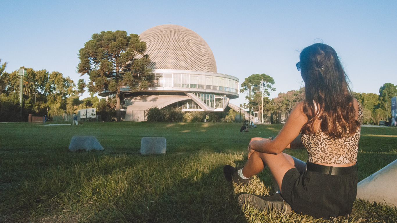 planetario-palermo Una mujer sentada sobre el césped contempla el icónico Planetario Galileo Galilei en Buenos Aires. La estructura futurista y el cielo despejado crean un escenario impresionante.