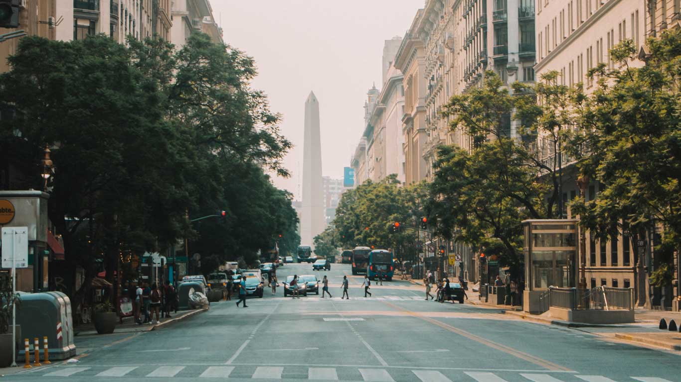 Avenida que conduce al Obelisco de Buenos Aires, un monumento icónico de Argentina y uno de los mejores planes para hacer en Buenos Aires. La calle está flanqueada por edificios altos y árboles, y varias personas cruzan el paso de peatones, con tráfico ligero visible. El Obelisco se alza destacado al fondo.