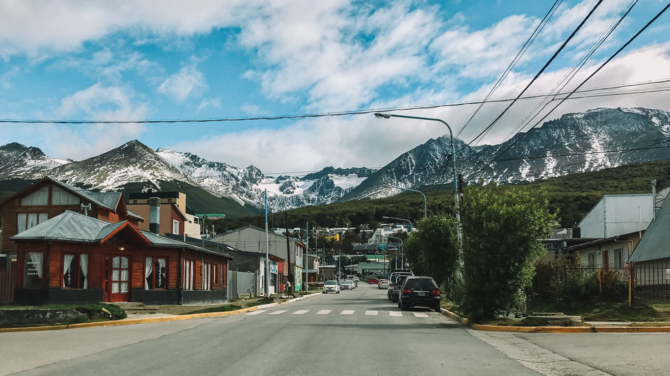 Calle tranquila en el centro, una de las mejores zonas donde alojarse en Ushuaia, con casitas de madera y coches aparcados, rodeada de montañas parcialmente nevadas bajo un cielo azul con nubes dispersas.