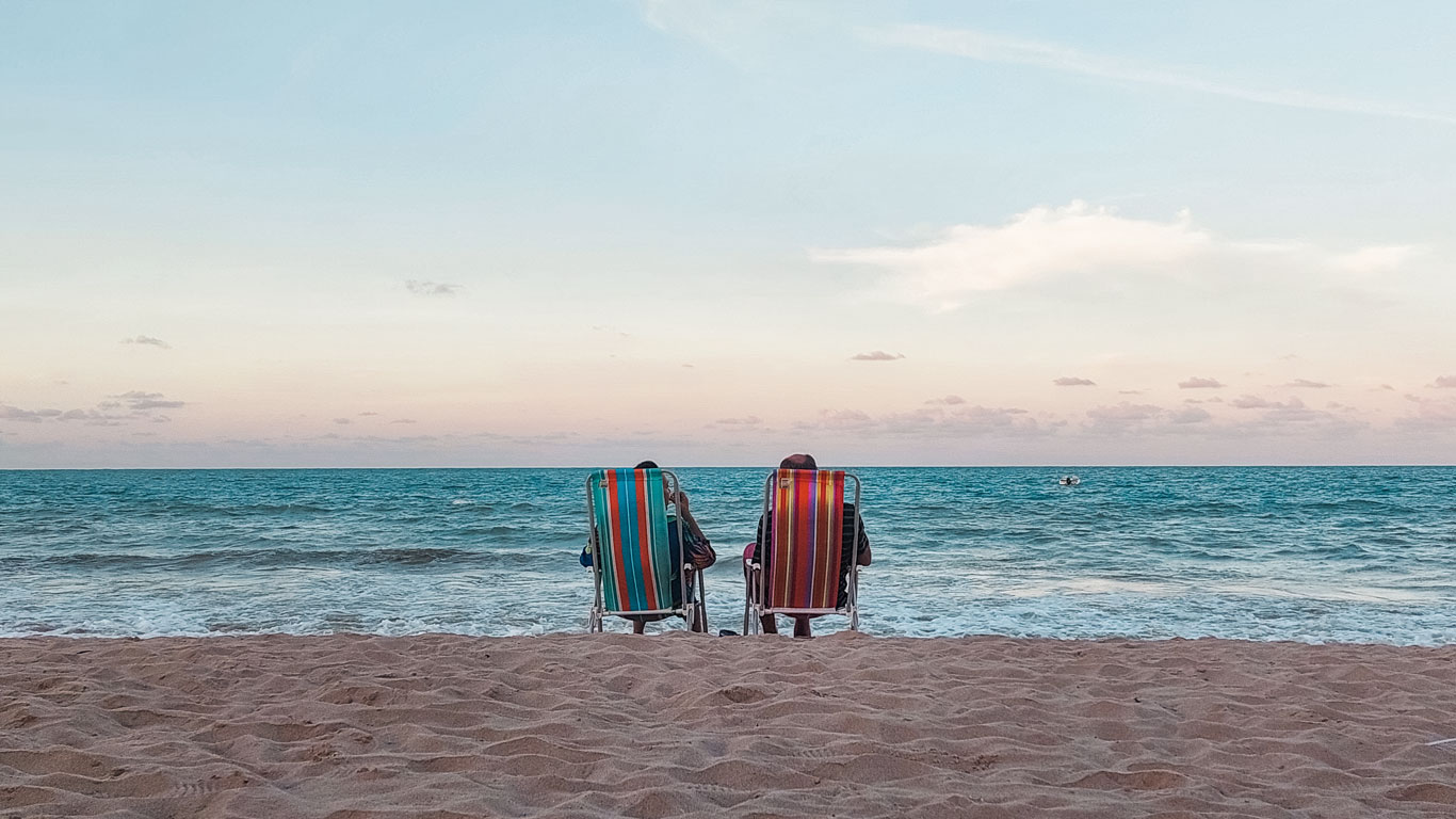 Dos personas sentadas en sillas de playa coloridas, de cara al mar tranquilo al atardecer en la Playa de Bessa, con el cielo en tonos pastel.