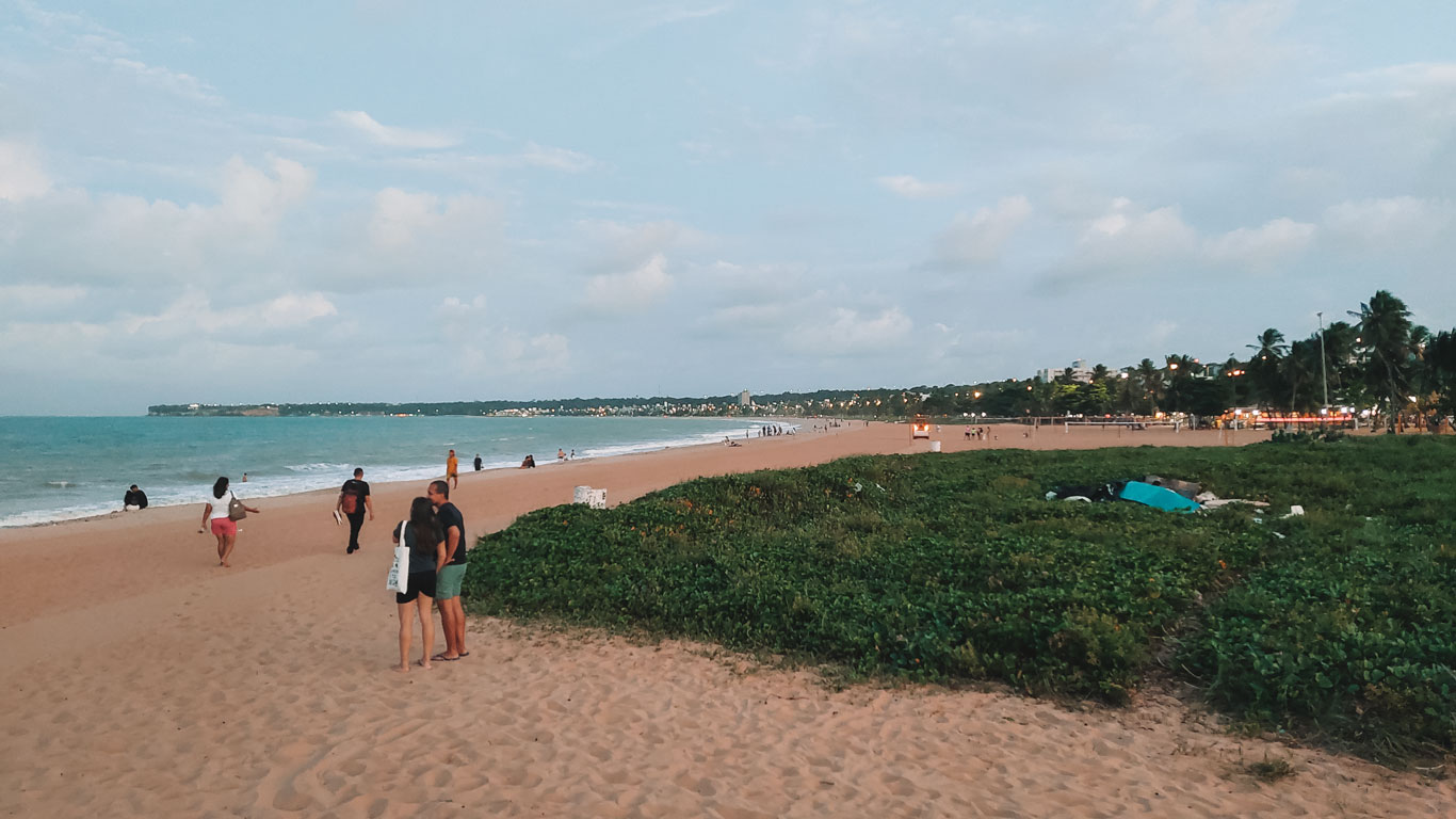 Personas caminando y charlando por la orilla de la Playa de Cabo Branco, con vegetación baja a la derecha y la ciudad de João Pessoa al fondo.