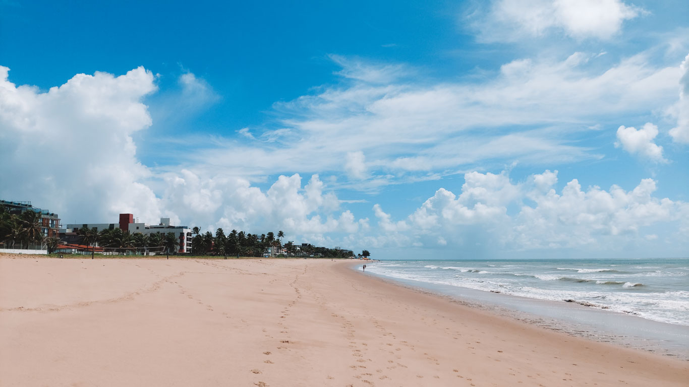 Playa de Jardim Oceania, sin bañistas, con arena clara y olas suaves, bajo un cielo azul con nubes esponjosas. Edificios bajos y palmeras a la izquierda, en João Pessoa.