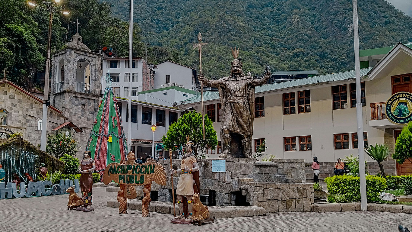 Estatua inca en la plaza principal de Aguas Calientes, rodeada de construcciones turísticas y vegetación. Este es el corazón de donde alojarse en Machu Picchu, con alojamientos, restaurantes y fácil acceso al autobús que lleva a las ruinas.