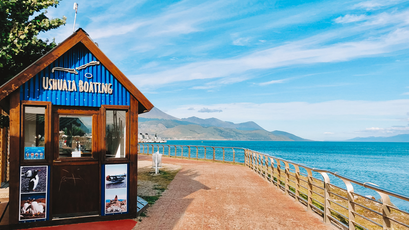 Pequeño chiringuito de madera con tejado azul y el cartel “Ushuaia Boating”, frente a un paseo con vistas a las montañas y al Canal Beagle.