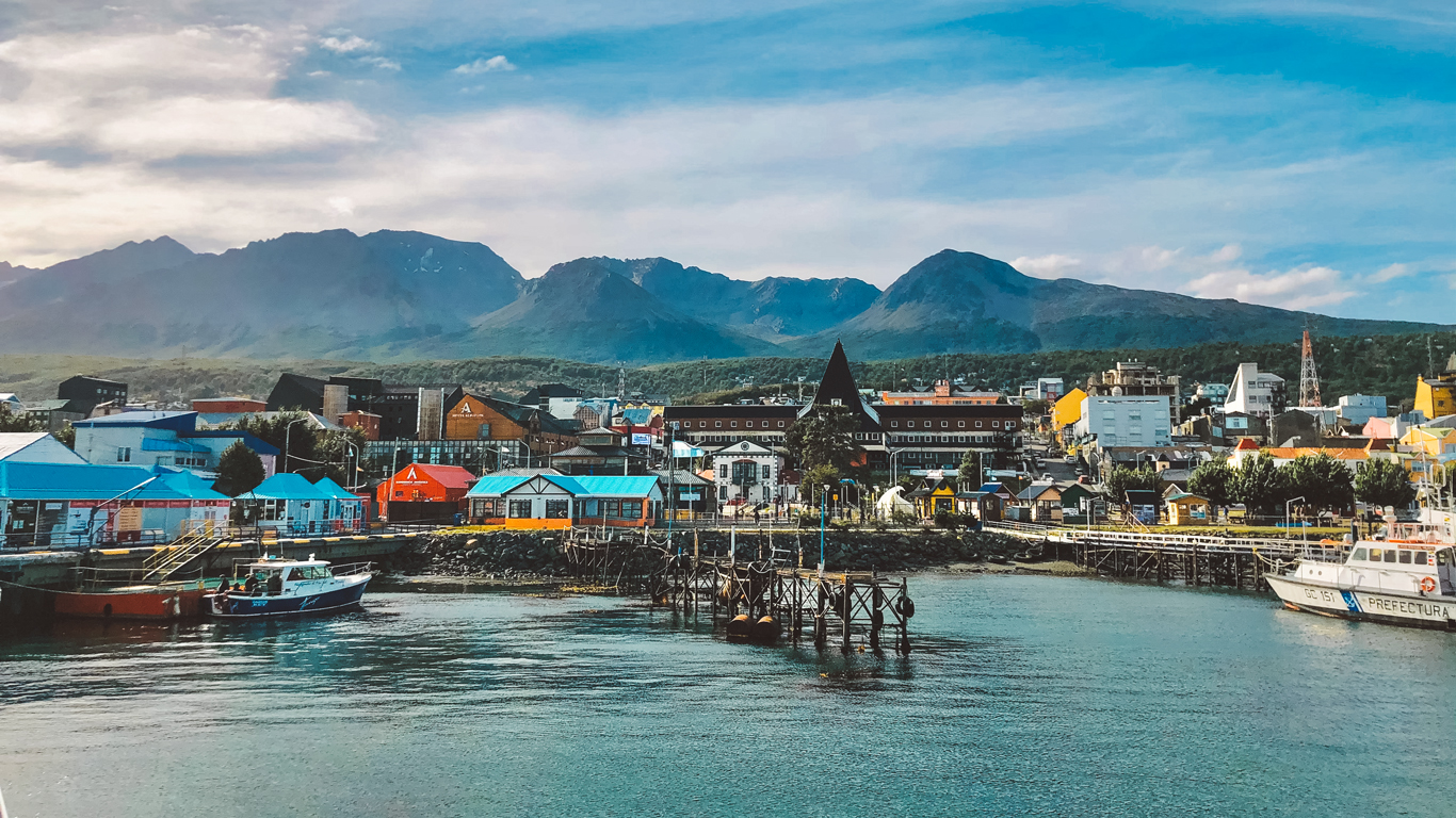 Vista del puerto de Ushuaia con casas de colores, barcos atracados y montañas al fondo; el cielo parcialmente nublado resalta la arquitectura con encanto de la ciudad.