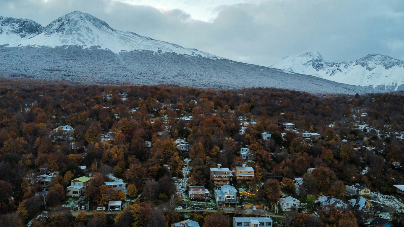 Vista aérea de un barrio residencial cerca del centro de Ushuaia, rodeado por un bosque con árboles en tonos otoñales, y con montañas nevadas al fondo dominando el paisaje.