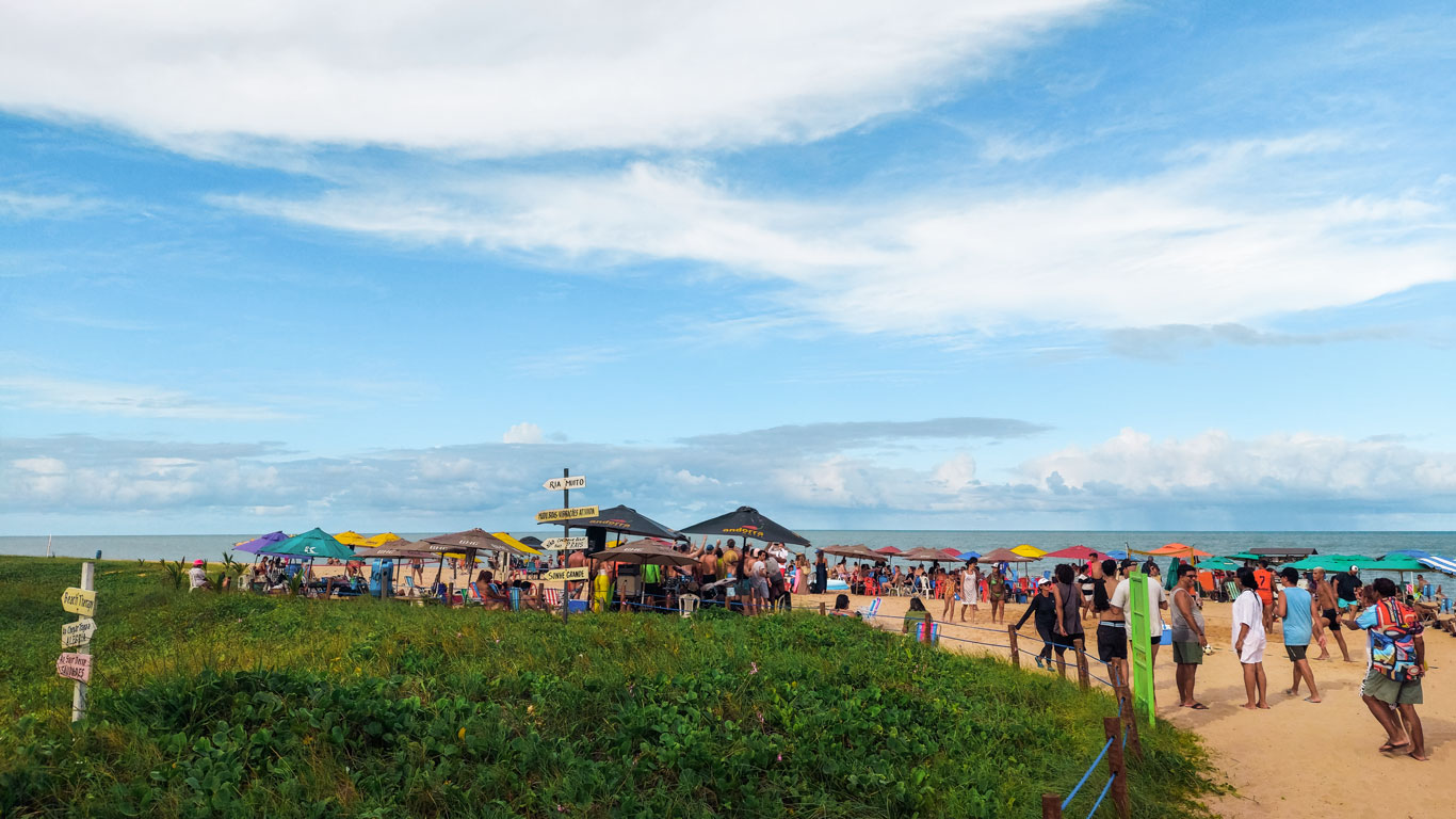 Playa concurrida de Jardim Oceania, con varias casetas coloridas y gente paseando, junto a una zona de césped con señales que indican direcciones.
