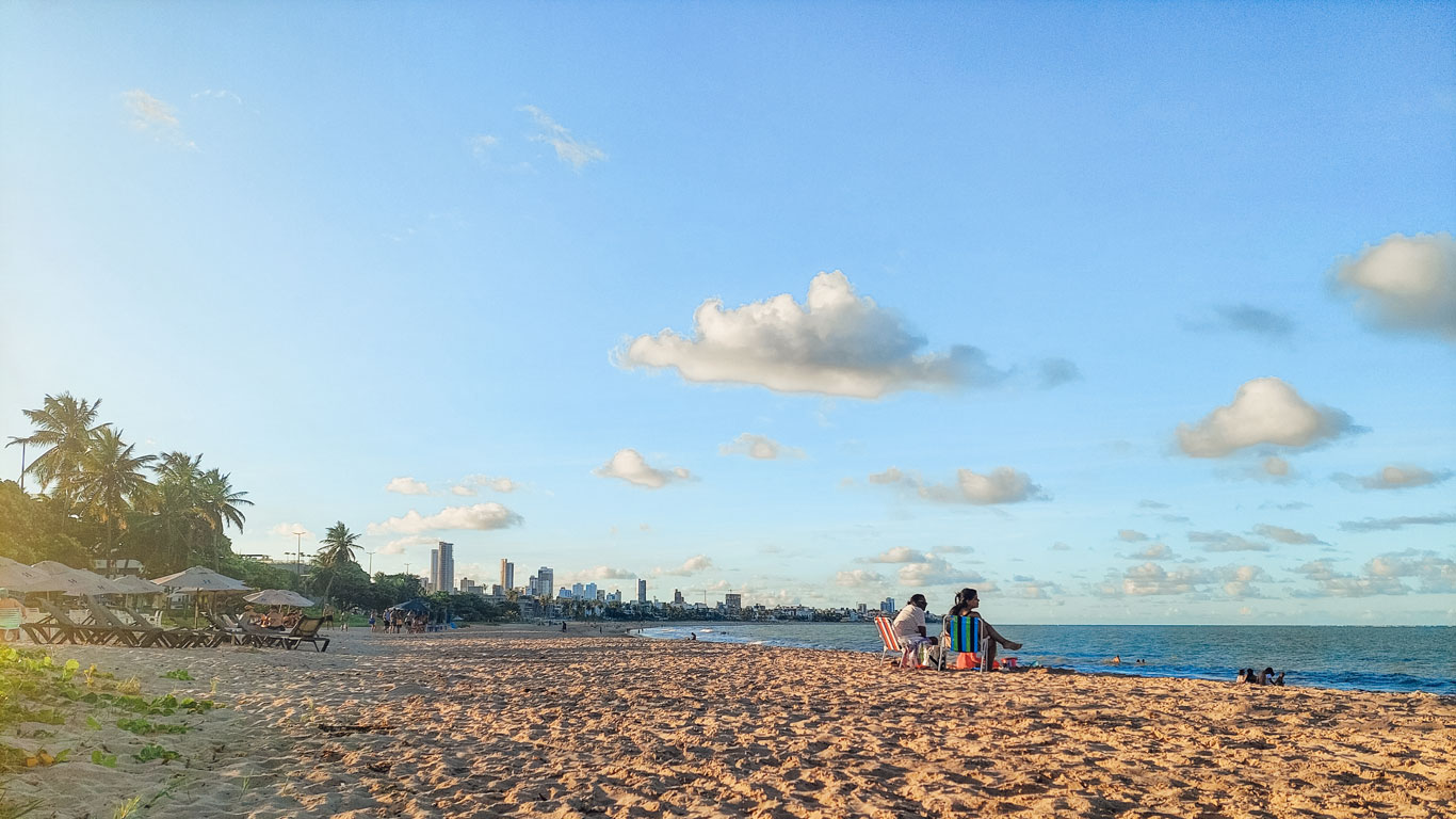 Playa de Manaíra con palmeras y sillas vacías a la izquierda, mientras algunas personas descansan en la arena a la derecha. Al fondo, el skyline de João Pessoa bajo un cielo azul dorado del final de la tarde.