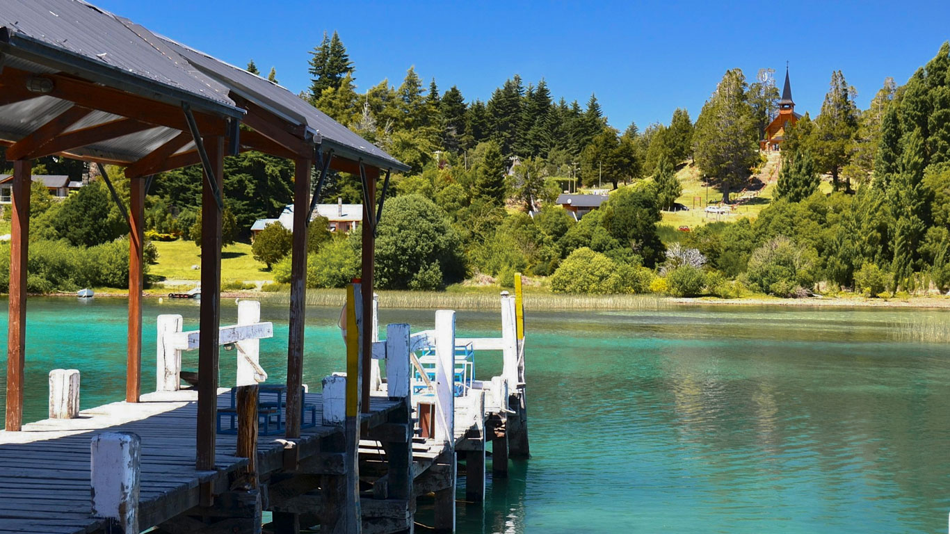 Antiguo muelle de madera a orillas de un lago cristalino en Bariloche, con aguas verde esmeralda, rodeado de exuberante vegetación. En lo alto de la colina, una pequeña iglesia con torre puntiaguda destaca entre los árboles.