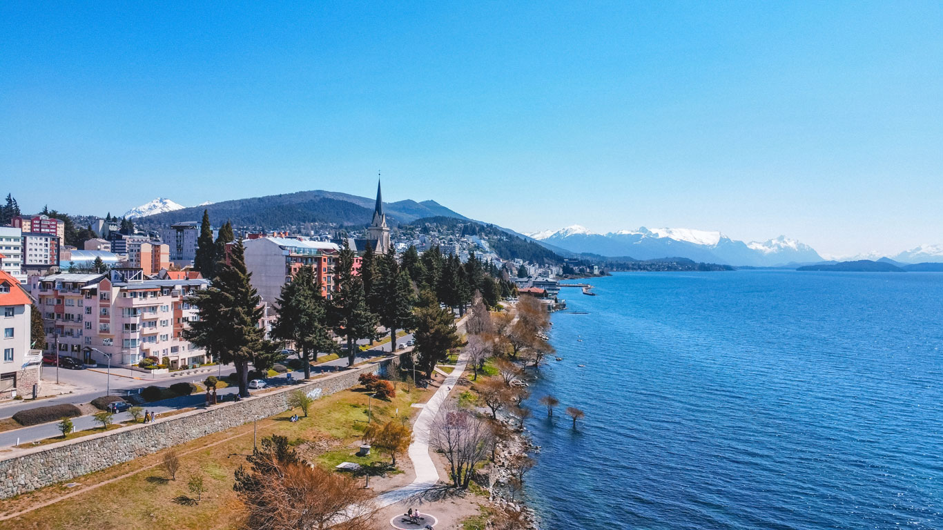 Vista aérea del centro de Bariloche, con edificios coloridos, una iglesia de torre alta y un paseo peatonal a orillas del Lago Nahuel Huapi, en el Centro de Bariloche, con montañas nevadas al fondo.