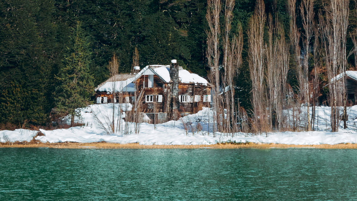 Cabaña de madera cubierta de nieve, rodeada de árboles altos y densos, frente a un lago de aguas verdosas en Bariloche.