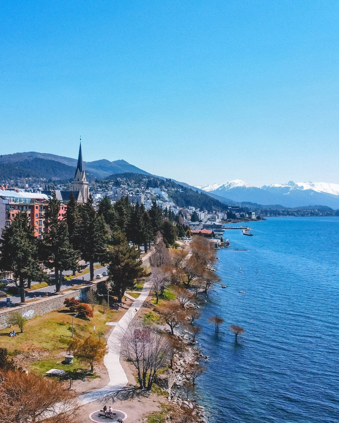 Un paseo a orillas de un lago cristalino en un día soleado, en pleno centro, el mejor lugar dónde alojarse en Bariloche. Con árboles dispersos, una iglesia con torre alta al fondo, y montañas nevadas en el horizonte. 