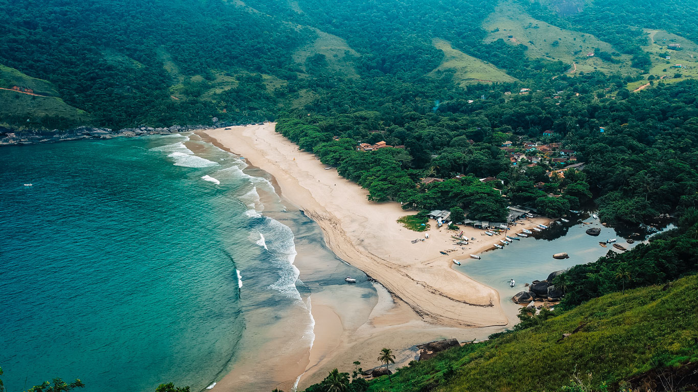 Imagen aérea de Praia do Bonete: el mar azul turquesa a la izquierda, una franja de arena clara en el centro y un pequeño poblado rodeado de vegetación densa a la derecha. Varias embarcaciones están ancladas en una ensenada tranquila, lo que convierte este rincón en un lugar ideal para quienes buscan alojarse en Ilhabela con acceso directo a la naturaleza y al mar.