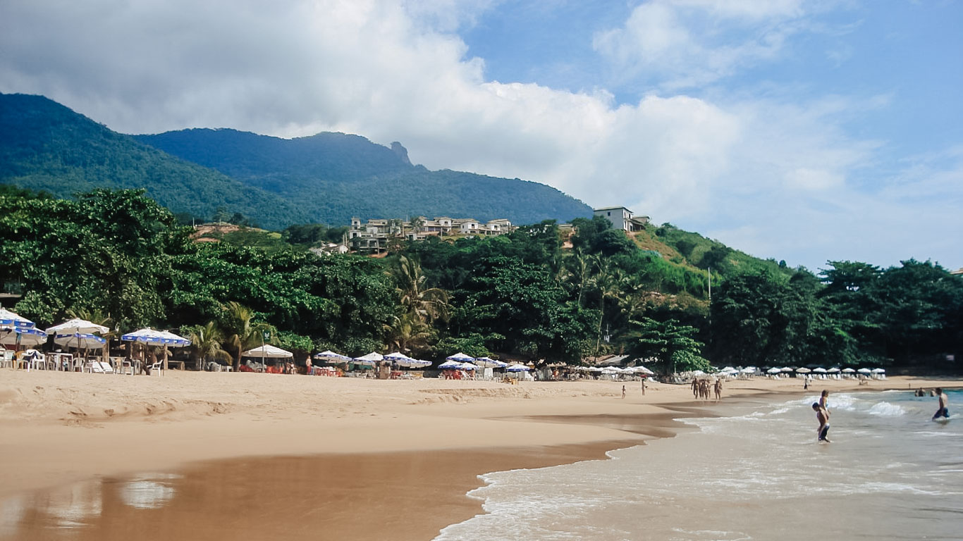 Praia do Curral, en Ilhabela, es uno de los mejores lugares donde alojarse en la isla. Su arena dorada, el mar tranquilo y las sombrillas alineadas a lo largo de la playa crean un ambiente perfecto para disfrutar del día. Al fondo, las colinas cubiertas por la Mata Atlántica y las casas en la ladera ofrecen vistas privilegiadas al mar.
