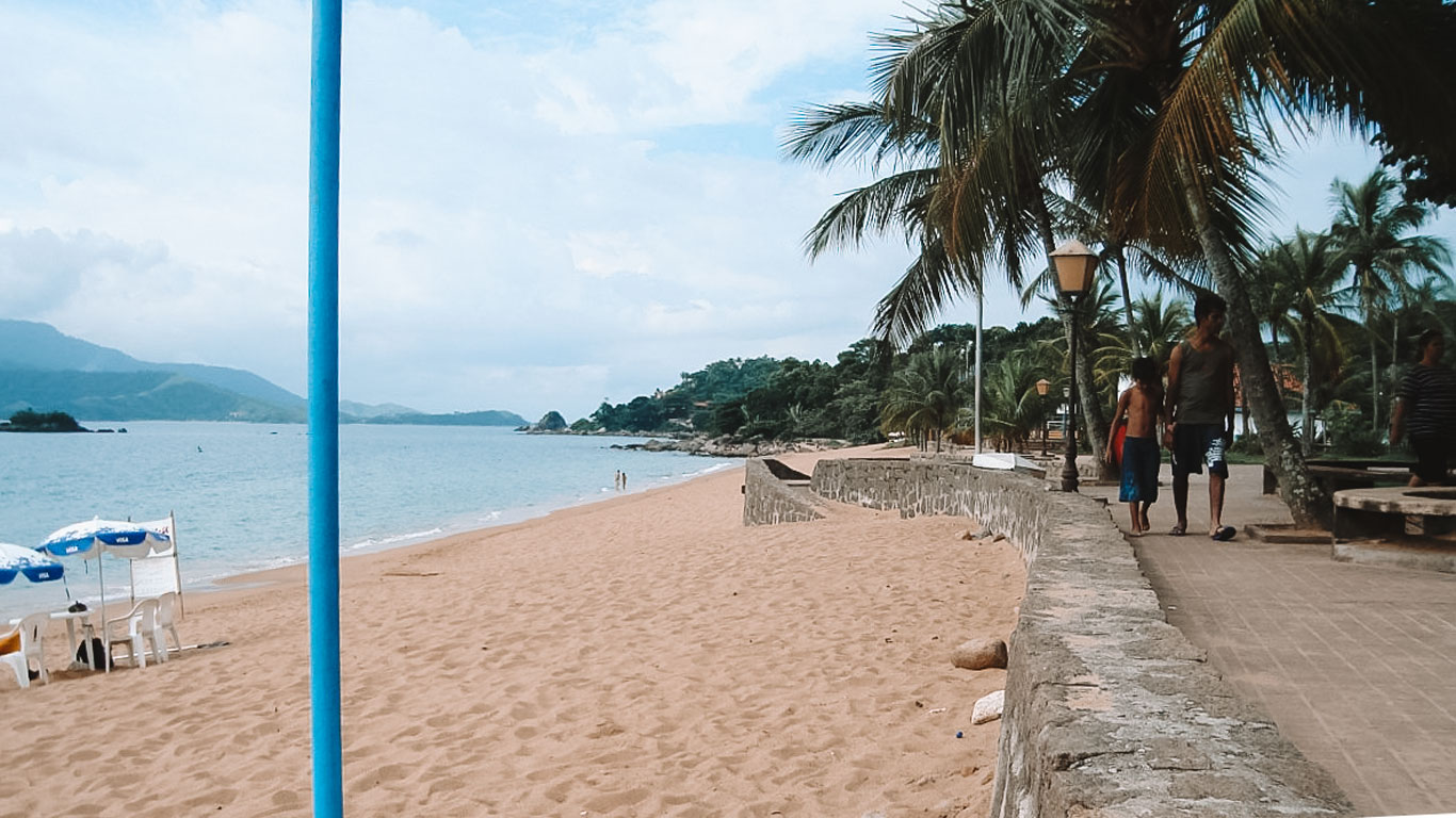 Paseo marítimo en Praia Grande, Ilhabela, con palmeras alineadas, bancos de piedra y gente paseando. A la izquierda, la franja de arena clara se extiende hasta el mar tranquilo, donde hay sombrillas y sillas vacías, mientras que al fondo se alzan colinas cubiertas de vegetación.