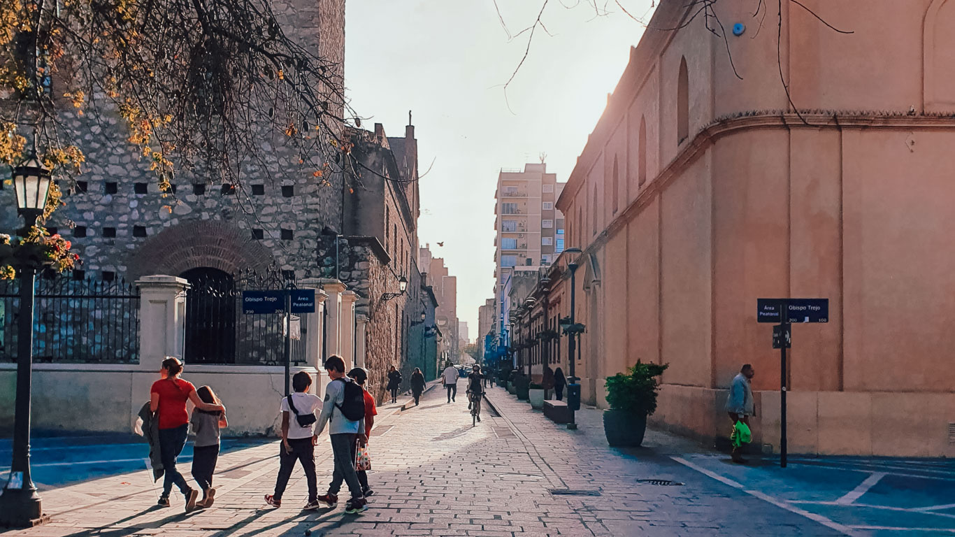 Personas caminando por una calle adoquinada en el centro histórico de Córdoba, Argentina, al atardecer. A la izquierda, un edificio de piedra antigua y a la derecha, construcciones coloniales iluminadas por la luz dorada del sol.