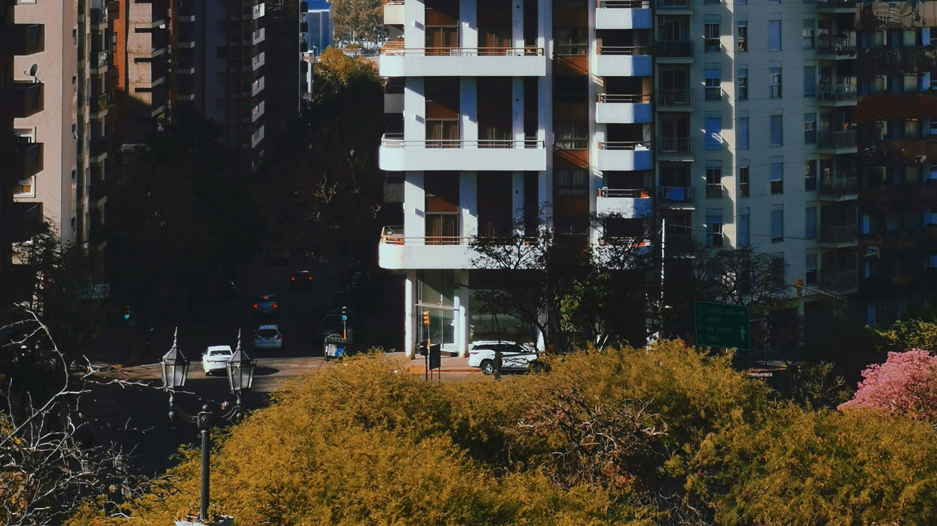 Imagen de edificios residenciales altos en una calle sombreada de Córdoba, vista desde arriba, con árboles dorados en primer plano. Al fondo, coches circulan por la calle entre los edificios.
