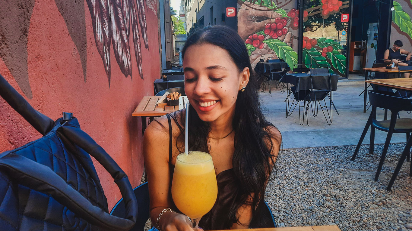 Mujer sonriente sosteniendo un vaso grande de zumo amarillo con pajita, sentada en una cafetería al aire libre con murales coloridos en las paredes, en Güemes, una de las mejores zonas donde alojarse en Córdoba, Argentina. El ambiente tiene mesas vacías y una decoración urbana con arte relacionado con el café y hojas.
