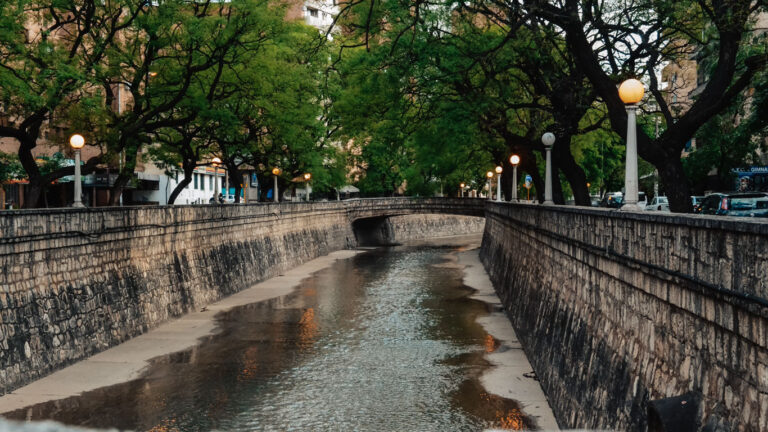 Vista del canal La Cañada en Córdoba con poca agua fluyendo, bordeado por muros de piedra y árboles verdes. Farolas acompañan las aceras a ambos lados del canal.