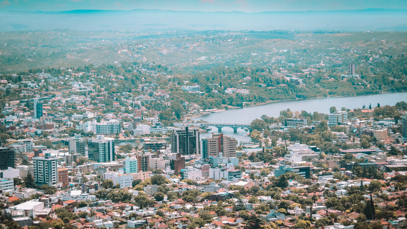 Panorama de Villa Carlos Paz con numerosos edificios, casas y zonas verdes que se extienden hasta las montañas al fondo. El río San Antonio atraviesa la ciudad, con un puente visible en el centro de la imagen.