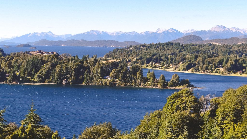 Vista panorámica de un lago azul profundo rodeado de densos bosques y montañas nevadas al fondo en Bariloche en noviembre, Argentina.