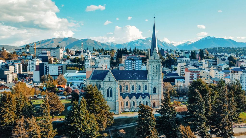 La Catedral de Bariloche, de estilo gótico, destaca con su alto campanario, rodeada de árboles y con la ciudad y las montañas al fondo. El cielo azul con nubes y la vegetación otoñal completan el paisaje urbano.
