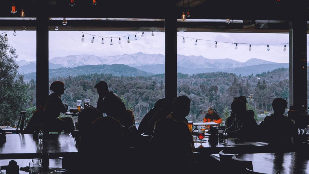 Interior de un restaurante acogedor con grandes ventanales panorámicos, donde personas charlan y disfrutan de bebidas mientras contemplan unas vistas impresionantes de las montañas y bosques de Bariloche en noviembre.