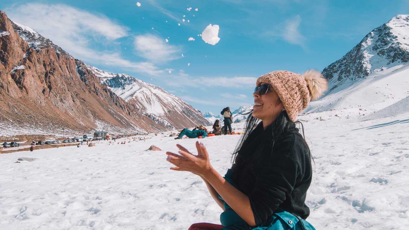 Mujer sonriente, con gorro de lana con pompón y gafas de sol, juega lanzando nieve al aire durante una excursión invernal en la Alta Montaña de Mendoza. Al fondo, montañas parcialmente cubiertas de nieve y un cielo azul claro de invierno, una buena época para quienes buscan cuándo ir a Mendoza para ver nieve.