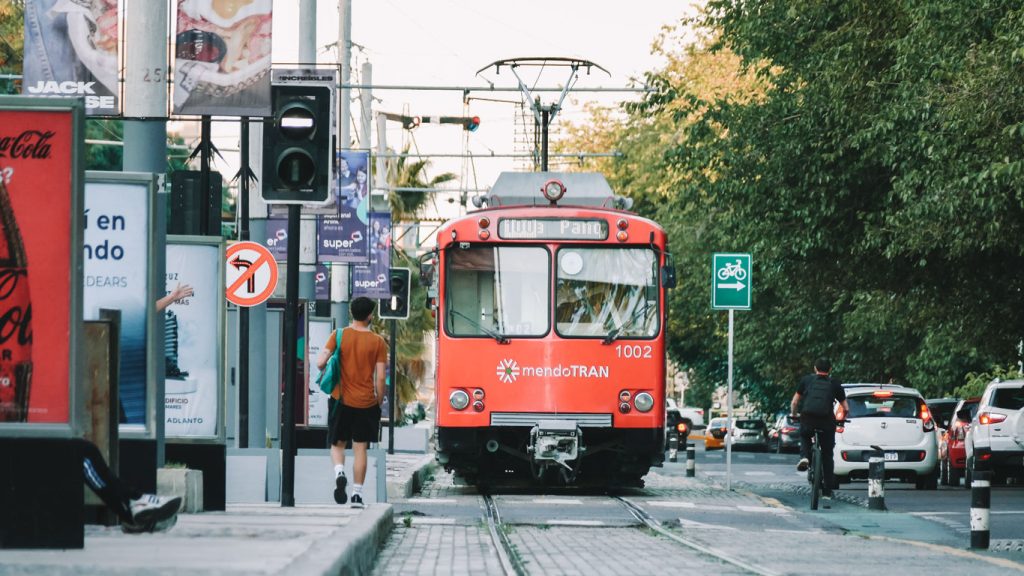 Tranvía rojo de la línea "mendoTRAN" circulando por las calles de Mendoza, rodeado de ciclistas, peatones y coches. Señales de tráfico, vallas publicitarias y árboles bordean la vía.
