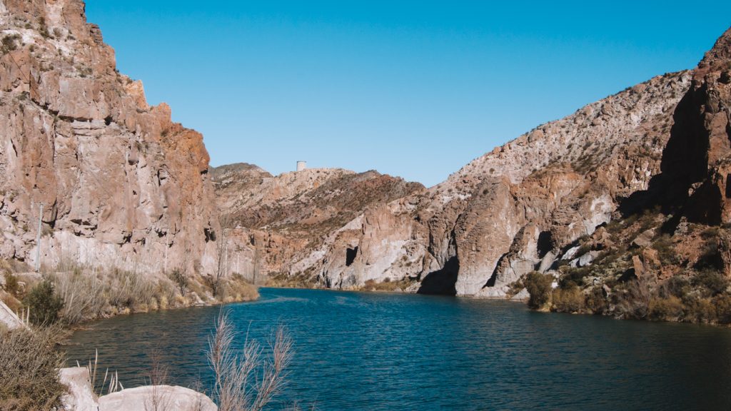 Cañón del Atuel con aguas azul oscuro que serpentean entre montañas áridas y empinadas de rocas rojizas en Mendoza. Vegetación baja crece a orillas del río bajo un cielo despejado.
